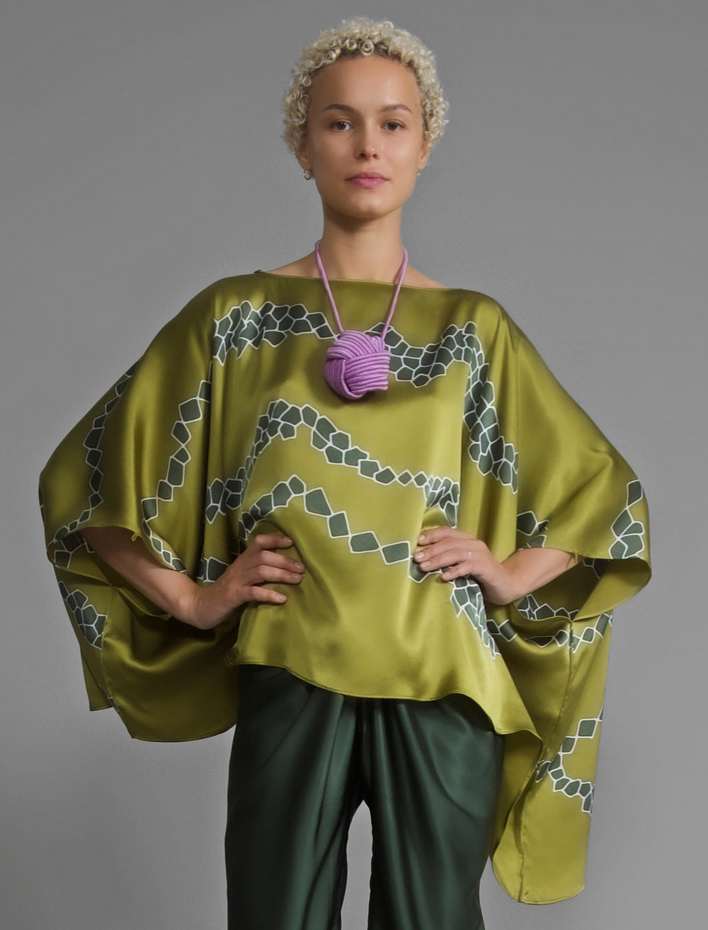 Front view. Flowing moss green colour silk top with graphic Macrosnake batik motif in contrasting green tone, featuring fluid draped sleeves, asymmetrical hemline, and versatile contemporary silhouette designed to be worn three ways.