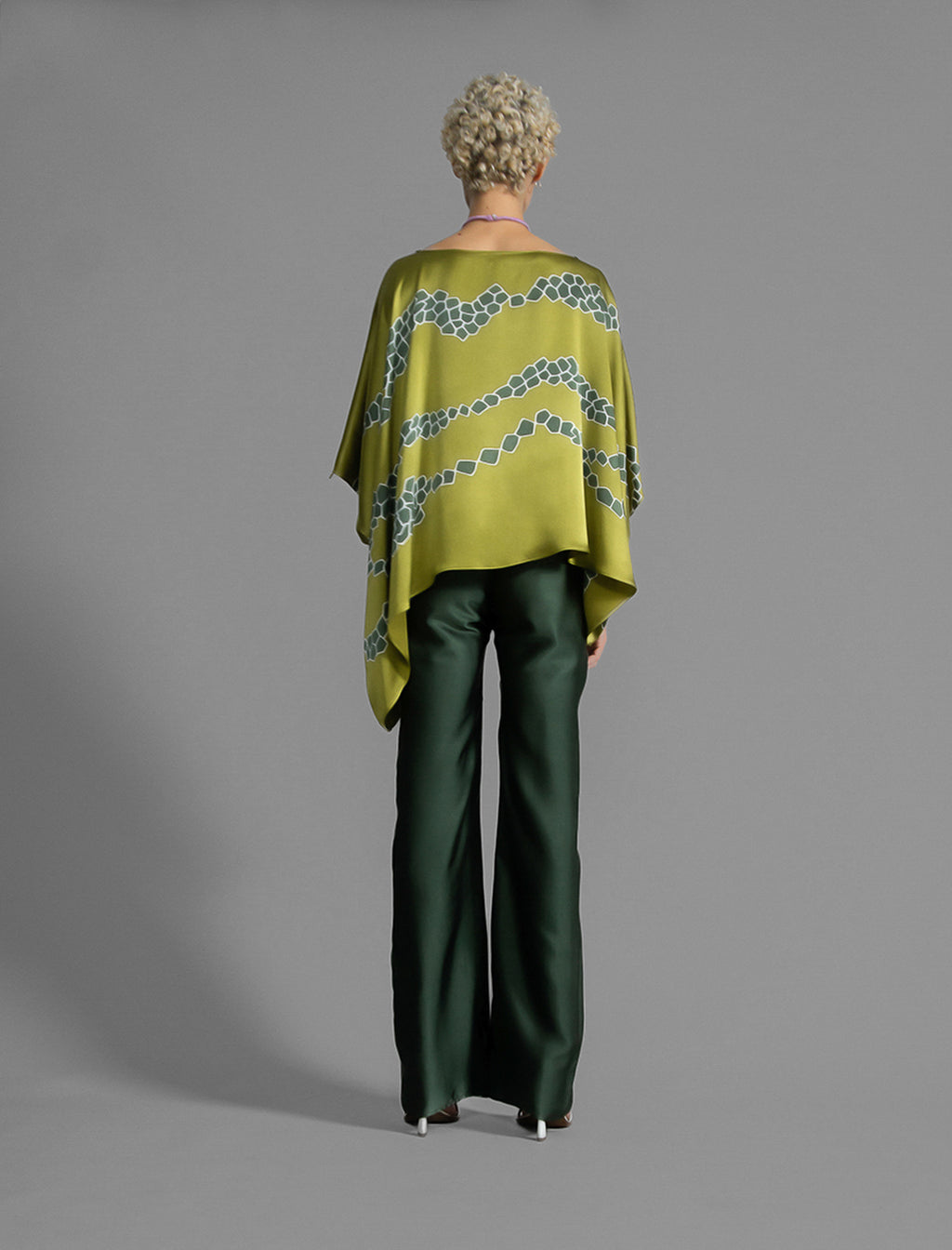 Back view. Flowing moss green colour silk top with graphic Macrosnake batik motif in contrasting green tone, featuring fluid draped sleeves, asymmetrical hemline, and versatile contemporary silhouette designed to be worn three ways.