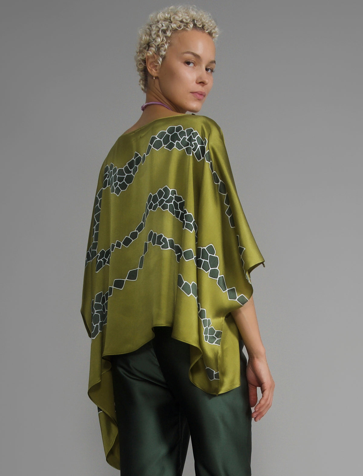Back view. Flowing moss green colour silk top with graphic Macrosnake batik motif in contrasting green tone, featuring fluid draped sleeves, asymmetrical hemline, and versatile contemporary silhouette designed to be worn three ways.