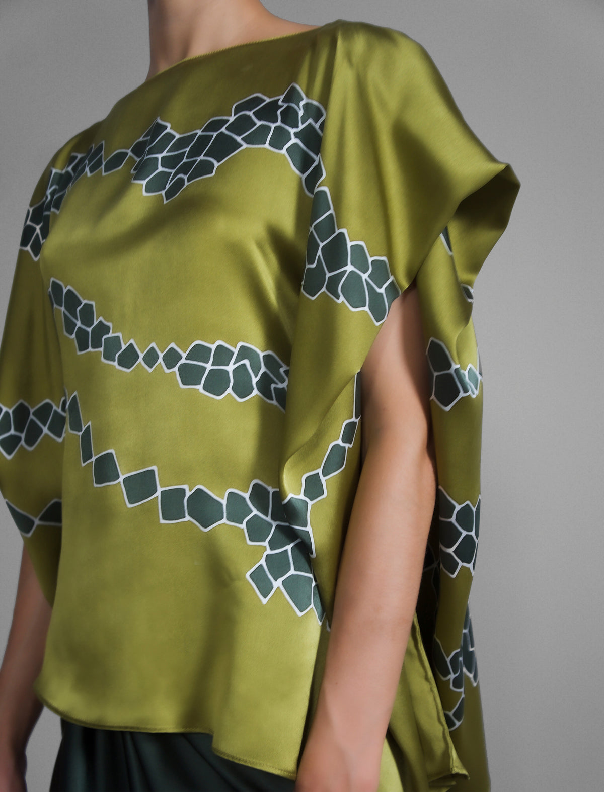 Short sleeve detail: Flowing moss green colour silk top with graphic Macrosnake batik motif in contrasting green tone, featuring fluid draped sleeves, asymmetrical hemline, and versatile contemporary silhouette designed to be worn three ways.