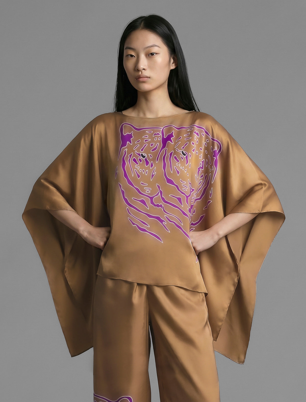 Front view. Flowing tobacco colour silk top with graphic Tiger batik motif in contrasting purple, featuring fluid draped sleeves, asymmetrical hemline, and versatile contemporary silhouette designed to be worn three wa