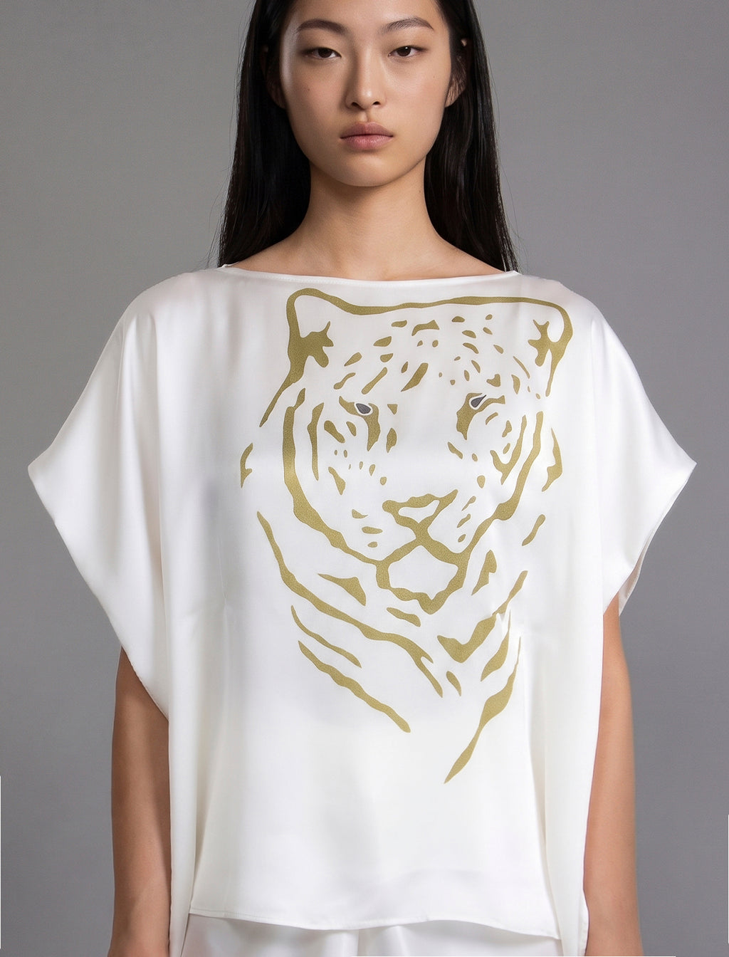 Front view, folded sleeve. Flowing white colour silk top with graphic Tiger batik motif in contrasting gold, featuring fluid draped sleeves, asymmetrical hemline, and versatile contemporary silhouette designed to be worn three ways.