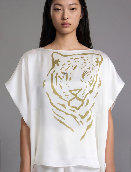 Front view, folded sleeve. Flowing white colour silk top with graphic Tiger batik motif in contrasting gold, featuring fluid draped sleeves, asymmetrical hemline, and versatile contemporary silhouette designed to be worn three ways.