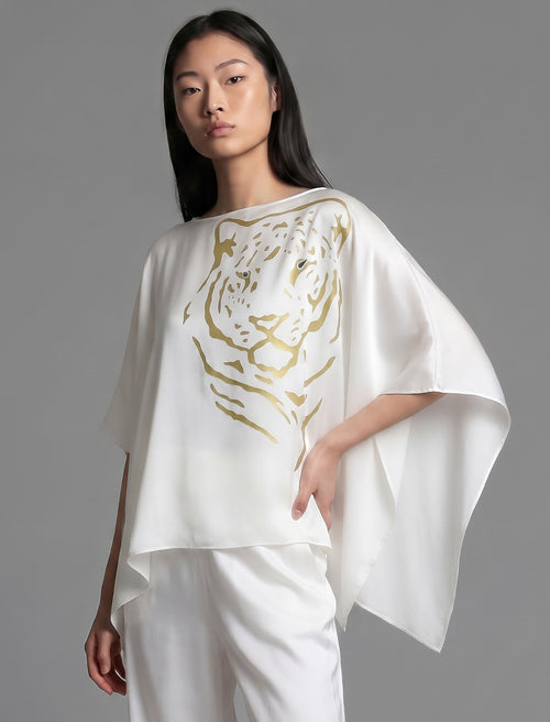 Front view, full sleeve. Flowing white colour silk top with graphic Tiger batik motif in contrasting gold, featuring fluid draped sleeves, asymmetrical hemline, and versatile contemporary silhouette designed to be worn three ways.