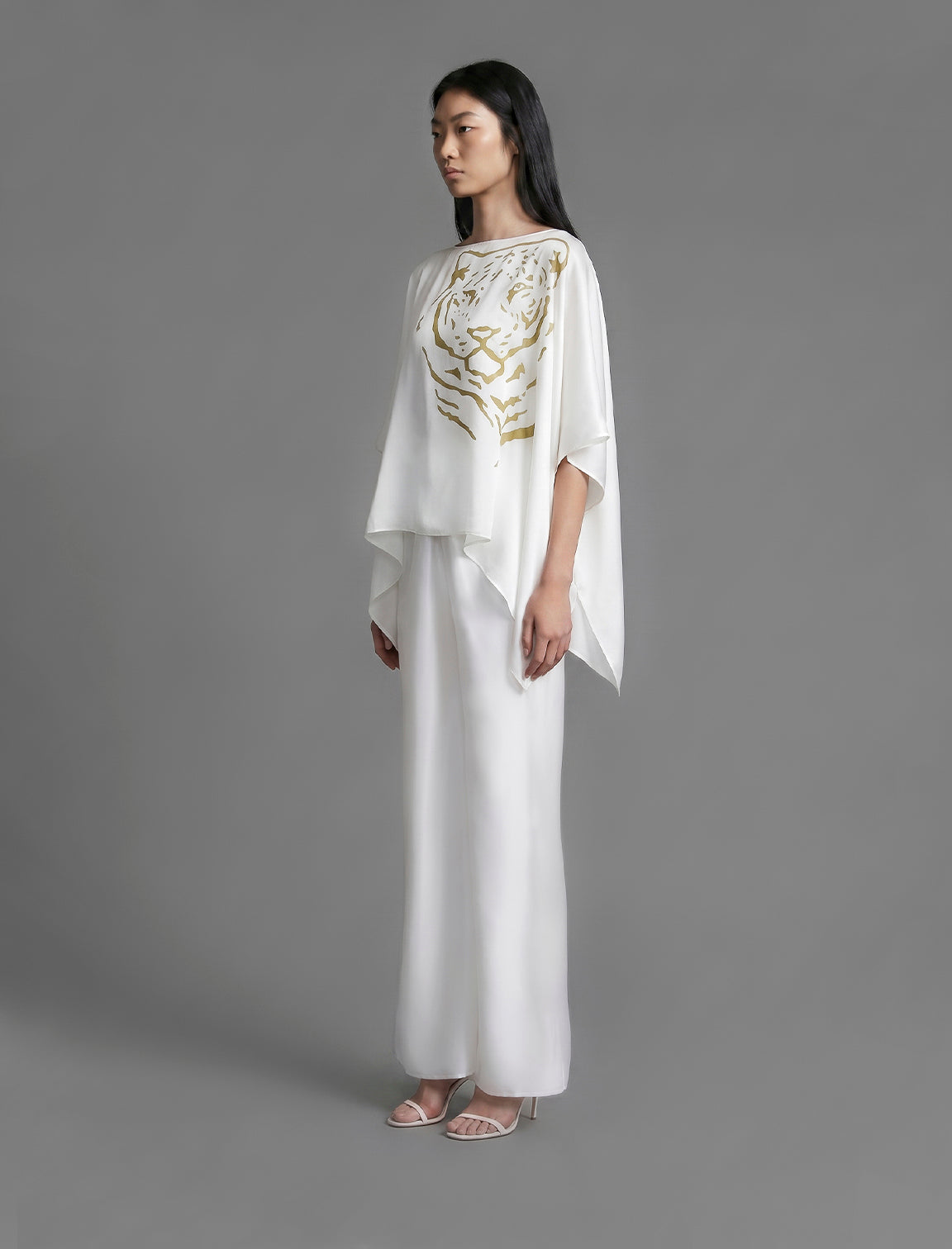 Full length view. Flowing white colour silk top with graphic Tiger batik motif in contrasting gold, featuring fluid draped sleeves, asymmetrical hemline, and versatile contemporary silhouette designed to be worn three ways.