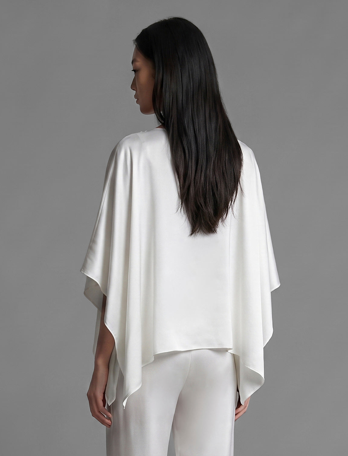 Back view. Flowing white colour silk top with graphic Tiger batik motif in contrasting gold, featuring fluid draped sleeves, asymmetrical hemline, and versatile contemporary silhouette designed to be worn three ways.