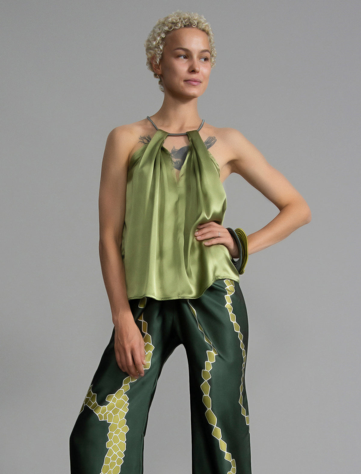 Front view. Silk satin halter top in fern green colour with elegant draping down the front, a high neckline anchored by a thin, grey silk cord that threads through the fabric, creating a V-shaped opening above the chest. Also draped at the back, the cord ties into a bow over a V-shaped opening. The hemline flows loosely over the hips.