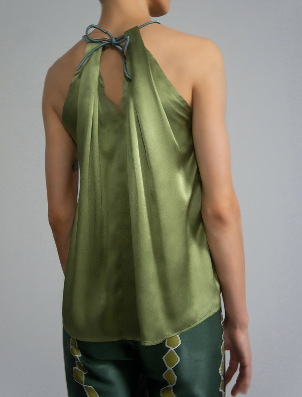 Back Detail: Silk satin halter top in fern green colour with elegant draping down the front, a high neckline anchored by a thin, grey silk cord that threads through the fabric, creating a V-shaped opening above the chest. Also draped at the back, the cord ties into a bow over a V-shaped opening. The hemline flows loosely over the hips.