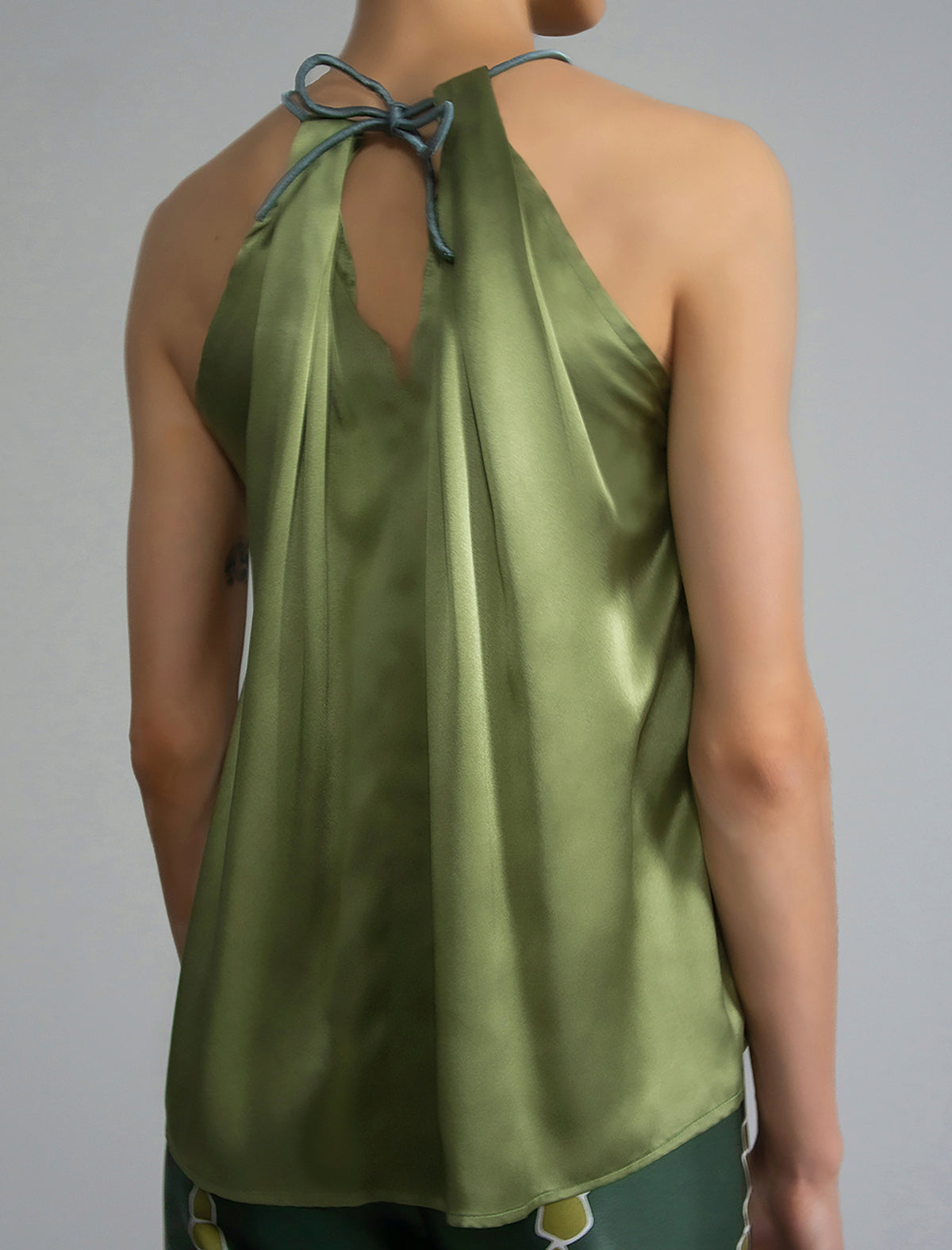 Back Detail: Silk satin halter top in fern green colour with elegant draping down the front, a high neckline anchored by a thin, grey silk cord that threads through the fabric, creating a V-shaped opening above the chest. Also draped at the back, the cord ties into a bow over a V-shaped opening. The hemline flows loosely over the hips.
