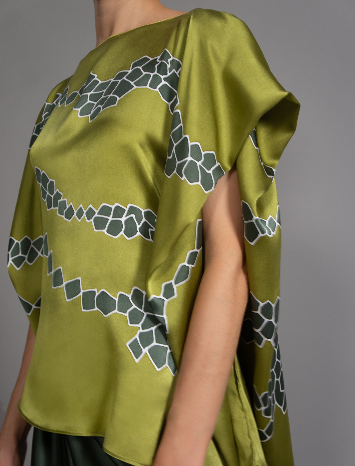 Short sleeve detail: Flowing moss green colour silk top with graphic Macrosnake batik motif in contrasting green tone, featuring fluid draped sleeves, asymmetrical hemline, and versatile contemporary silhouette designed to be worn three ways.