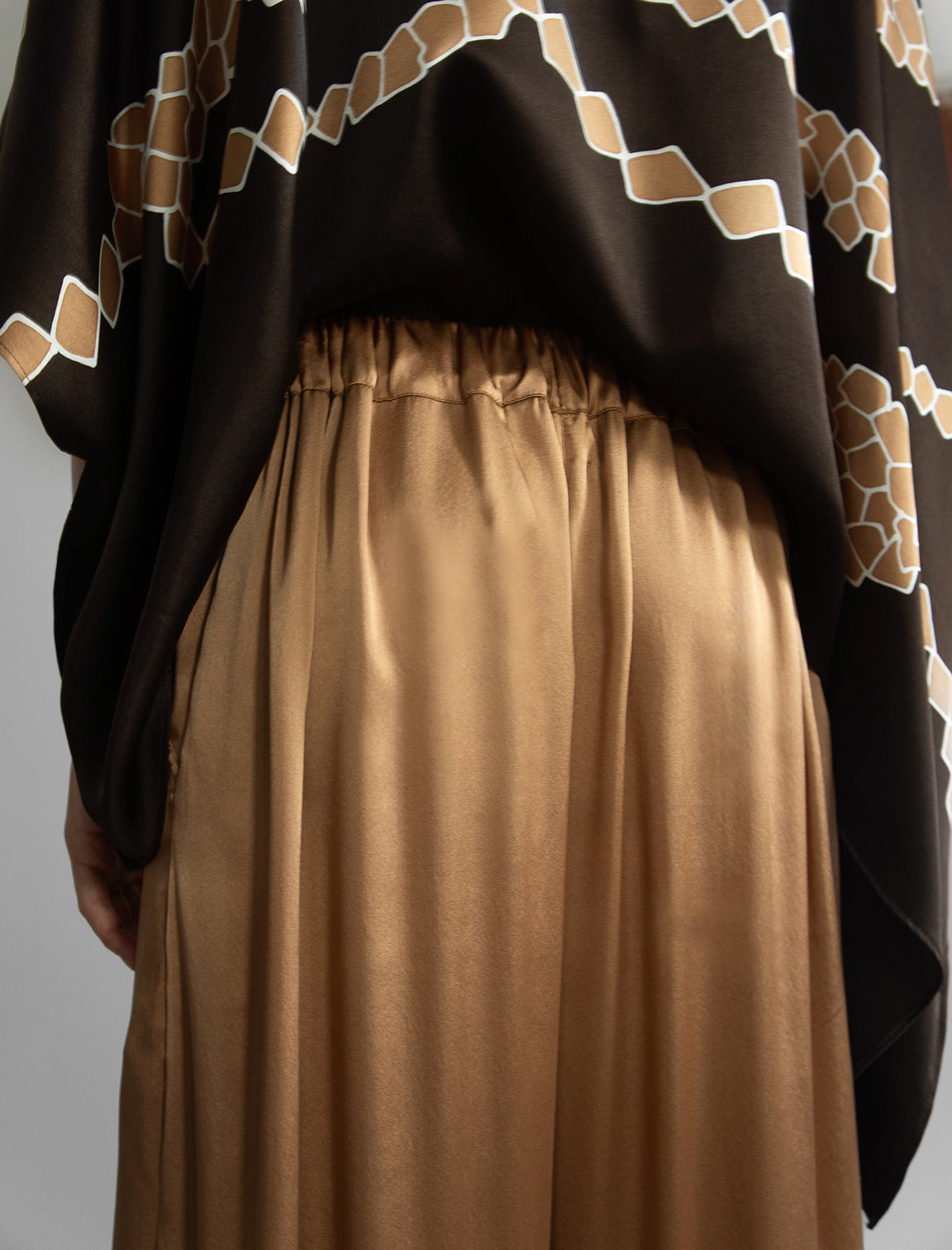 Gathered waistband detail: Relaxed wide-leg silk trousers in warm tobacco, featuring drawstring waistband, elasticated at the back. Soft gathered drape, fluid silhouette, and subtle lustrous finish.