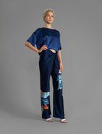 Right side view. Long silk pants in blue navy colour with oversized tropical leaf batik motif, featuring a knot tie at the front and a slick relaxed silhouette.