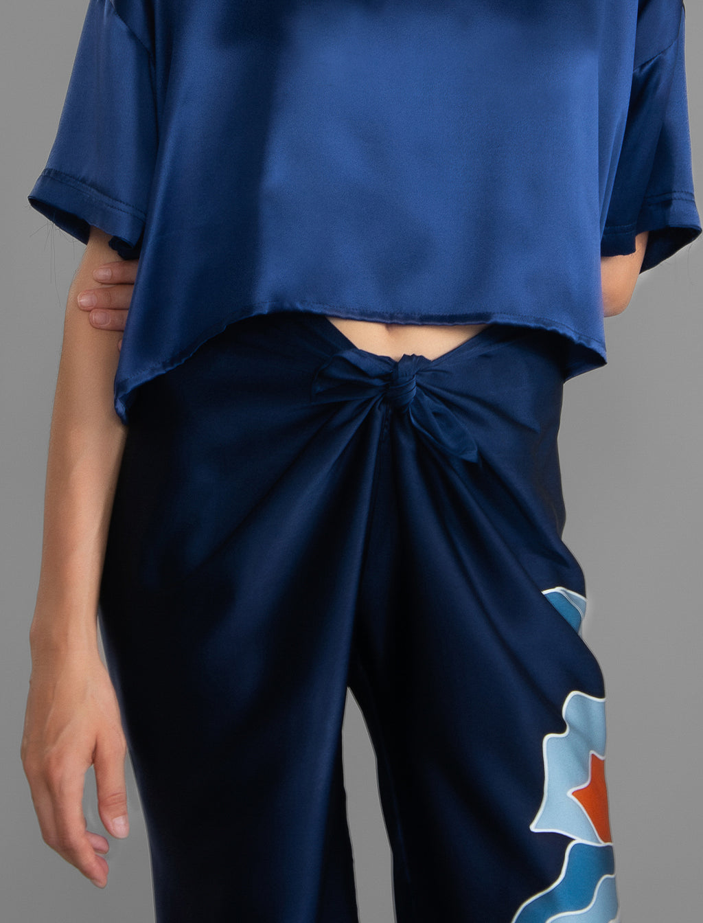 Knot detail view. Long silk pants in blue navy colour with oversized tropical leaf batik motif, featuring a knot tie at the front and a slick relaxed silhouette.