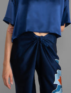 Knot detail view. Long silk pants in blue navy colour with oversized tropical leaf batik motif, featuring a knot tie at the front and a slick relaxed silhouette.