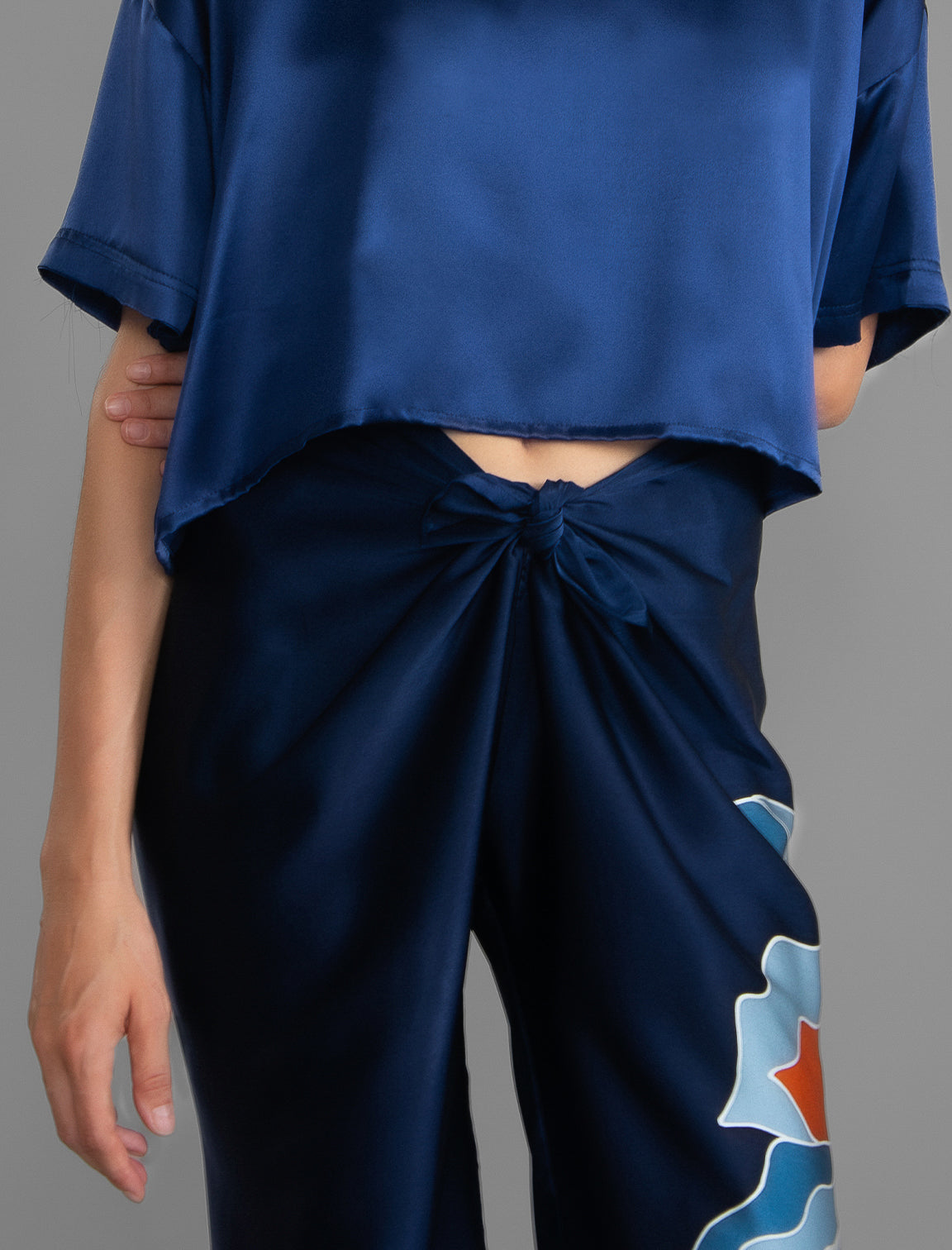 Knot detail view. Long silk pants in blue navy colour with oversized tropical leaf batik motif, featuring a knot tie at the front and a slick relaxed silhouette.