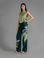 Side view. Long silk pants in teal colour with oversized tropical leaf batik motif, featuring a knot tie at the front and a slick relaxed silhouette.