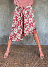 Load image into Gallery viewer, This asymmetric Skirt-Pant is handcrafted from silk satin and blur the line between shorts and skirt. A deep, gathered waistband flows into a fluid silhouette, finished with a diagonal hemline that brings an unconventional edge to refined dressing.This  hand-painted stamp batik motif takes inspiration from the late 19th-century ‘Wiener Secession’ art movement, infusing the design with a refined sense of modularity. Pink and ivory colors here are beautifully matched.


