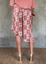 Load image into Gallery viewer, Back detail: this asymmetric Skirt-Pant is handcrafted from silk satin and blur the line between shorts and skirt. A deep, gathered waistband flows into a fluid silhouette, finished with a diagonal hemline that brings an unconventional edge to refined dressing.This  hand-painted stamp batik motif takes inspiration from the late 19th-century ‘Wiener Secession’ art movement, infusing the design with a refined sense of modularity. Pink and ivory colors here are beautifully matched.


