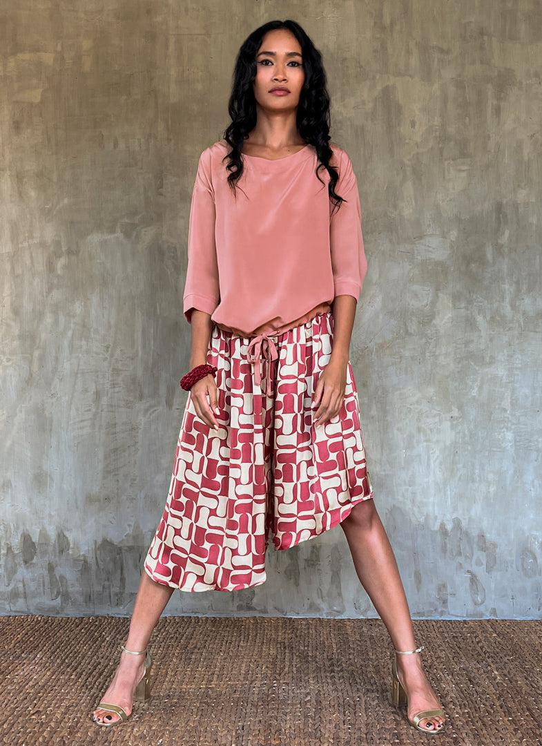 This asymmetric Skirt-Pant is handcrafted from silk satin and blur the line between shorts and skirt. A deep, gathered waistband flows into a fluid silhouette, finished with a diagonal hemline that brings an unconventional edge to refined dressing.This  hand-painted stamp batik motif takes inspiration from the late 19th-century ‘Wiener Secession’ art movement, infusing the design with a refined sense of modularity. Pink and ivory colors here are beautifully matched.