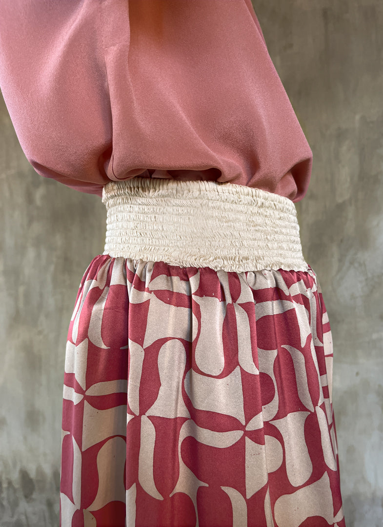Detail of elasticated waist: this asymmetric Skirt-Pant is handcrafted from silk satin and blur the line between shorts and skirt. A deep, gathered waistband flows into a fluid silhouette, finished with a diagonal hemline that brings an unconventional edge to refined dressing.This  hand-painted stamp batik motif takes inspiration from the late 19th-century ‘Wiener Secession’ art movement, infusing the design with a refined sense of modularity. Pink and ivory colors here are beautifully matched.