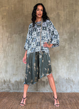 Load image into Gallery viewer, This asymmetric Skirt-Pant is handcrafted from silk satin and blur the line between shorts and skirt. A deep, gathered waistband flows into a fluid silhouette, finished with a diagonal hemline that brings an unconventional edge to refined dressing.This  hand-drawn batik motif is inspired from the late 19th-century ‘Wiener Secession’ art movement, infusing the design with a refined sense of linear rigor softened by small foliage details. here in steel grey background and beige motif.
