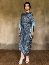 Load image into Gallery viewer, Asymmetric long Jogya silk dress with a &quot;Deco&quot; pattern in grey and dark blue colors. The dress features a loose fit with a belt and has a subtle asymmetrical design. Steel grey border define the kimono-like sleeves. Luxury resort-wear in silk batik made in Bali. 
