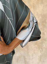 Load image into Gallery viewer, detail of 3/4-length split sleeves. One sleeve can be adjusted to two different lengths with a discreet button-and-loop fastening —Silk satin handpainted asymmetric long dress in Greenwall botanical pattern in light blue background and darker leaves .
