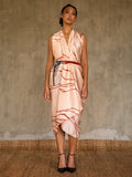 A woman wearing a pink champagne colored silk sarong with a geometric kintsugi pattern, tied in a knot at the waist, standing against a textured background.