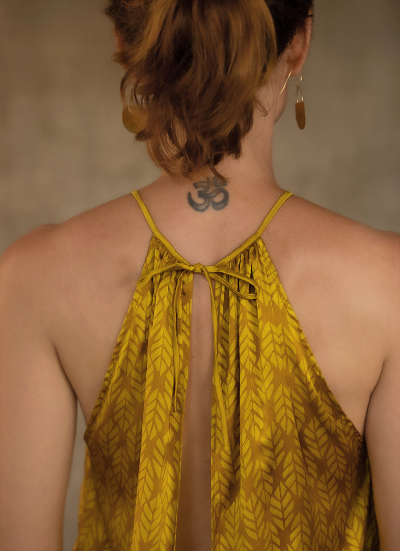 A female model wearing a satin silk halter top with a geometric leaf batik pattern in gold yellow and yellow color and a matching silk long pant grey with lines yellow motif produced in Bali